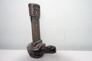 Vorbild ist eine Bixi-Skulptur aus der chinesische Mythologie (hier: Qing-Dynastie), so werden Mischwesen mit dem Körper einer Schildkröte und dem Kopf eines Drachen bezeichnet (Bixi ist einer der neun Söhne des Drachenkönigs). Ein optisch vergleichbares Objekt war ursprünglich ein Geschenk an Erzherzog Franz Ferdinand, der 1893 China besuchte. Es hatte die Funktion einer Petschaft, um ein Siegel einzudrücken. Um 1900 wurden dann solche Stücke in kleinerer Form (wie hier als Salzstreuer, befüllbar von unten) als Luxusgegenstände für den europäischen Markt hergestellt, Silber, Höhe 10 cm. Es gibt in ostasiatischen Länder wie China, Mongolai, Vietnam, Korea und Japan zum Teil große Skulpturen, u.a. mit Stelen, getragen von Schildkröten, aber natürlich auch kleine Exemplare für den privaten Bedarf (Suche: Bixi). Es soll Glück bringen, daran zu reiben. Der Ursprung liegt bereits im 3. Jhd. nach unserer Zeitrechnung.