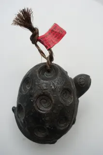Kleine Glocke aus Japan, gegossen und patiniert, unten offen, ein älteres Stück, mit dem geflochtenen Band 12 cm hoch. Das rote Fähnchen ist mit Glück- und Segenswünschen beschriftet. Experten gehen davon aus, dass es in China schon seit ca. 5.000 Jahren Glocken gibt. Der Glockenguss vereint handwerkliches Können, musikalisches Wissen und tief verwurzelte Traditionen.