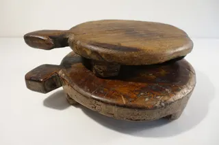 Zwei ältere Hocker, massives Tropenholz, in verwandter Form, der Kopf dient als Griff, beide aus jeweils einem Stück. Der untere kommt aus Ghana (dreibeinig, schon beschrieben), der obere aber aus Indien, wird dort als Chapattibrett bezeichnet, steht auf zwei Stegen und kann auch als Tischchen genutzt werden, Größe 29 cm.