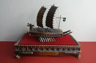 TURTLE SHIP ( sog. Schildkrötenschiff mit Drachenkopf/Bixi), Bronze, auf Metall-Sockel mit rotem Samt und Plexiglas-Sturz, 15,4 x 14 cm. Ein Souvenier, erhalten über den Künstler Duk Su Yun (weitere Infos unter "Suche : Duk Su Yun). Solche Schiffe wurden im Auftrag des Admirals Yi Sun-sin gebaut und erstmals 1592 gegen Japan von ihm eingesetzt. Er schlug in mehreren Seeschlachten die zahlenmäßig überlegene japanische Flotte, worauf man in Korea heute noch stolz ist. Ein originalgetreuer und sehr stattlicher Nachbau, aber u.a. auch Gedenkmedaillien und Briefmarken sind in Seoul im Museum zu sehen. Nach diesen Kriegen wurden größere Schildkrötenschiffe gebaut und mit einer Panzerung gegen Brandpfeile ausgestattet. Durch das Maul konnte man eine von insgesamt 30 Kanonen abfeuern. In meiner Sammlung befinden sich auch ein Geldschein und eine kleine Münze mit diesem Motiv. Ähnliche und größere Nachbauten dieses Schiffstyps konnte ich auf der Documenta 11 in Kassel (von Allan Sekula) sehen bzw. im Maritimen Museum Hamburg fotografieren.