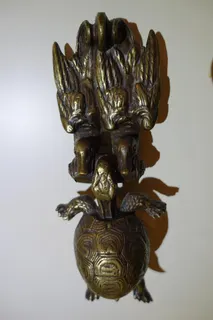 Türklopfer, Bronzeguss, Größe 21 cm. Die Schildkröte hängt mit dem Maul an einem Stock (gleichzeitig Scharnier), den zwei Enten im Schnabel tragen. Nach einer Fabel erfüllt sich so ihr Wunsch, fliegen zu können. Aber sie stürzt ab, weil sie vor Begeisterung jubelnd das Maul aufmacht. Die Moral von der Geschichte darf jeder selbst ziehen.